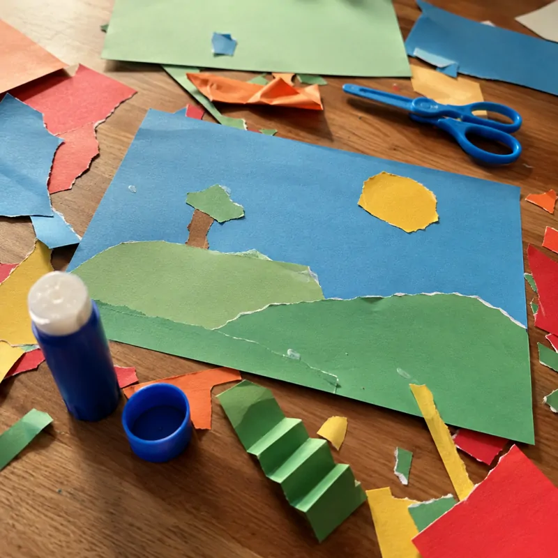 Collage de un paisaje con papel rasgado: un sol, una colina y un árbol, rodeado de recortes de papel y un pegamento en barra